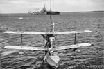A Supermarine Walrus amphibious aircraft is hoisted on board HMS Warspite after a sweep in the Indian Ocean. Crew members stand on both the top and the bottom wing steadying the aircraft during the lift. An Illustrious class aircraft carrier can be seen moored in the background. Alongside her is another vessel, possibly a hospital ship, and HMS Ludlow A cruiser is moored aft of these vessels and a motor cutter is making its way from this group of ships towards the camera. Photo: Lt. D. C. Ouids. Imperial War Museums A 10649